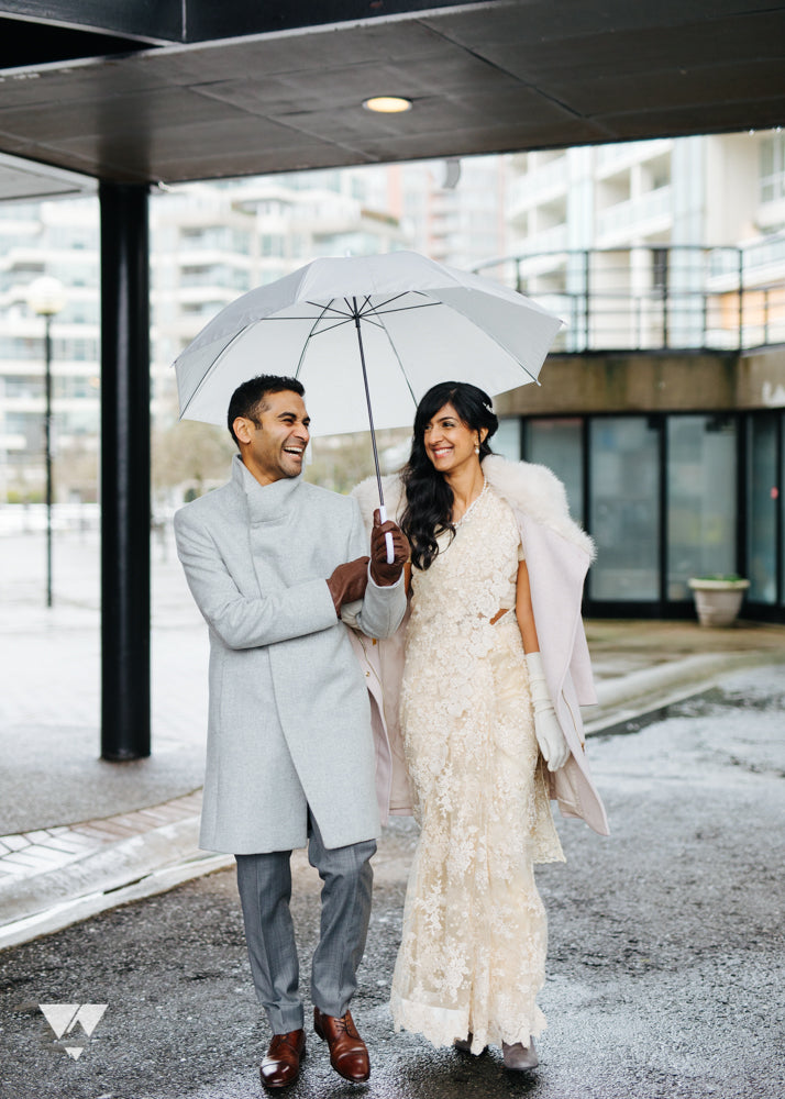 A couple walking outdoors under a white umbrella on a rainy day. The man wears a light gray overcoat and gray trousers, while the woman wears a cream lace wedding gown with a white faux-fur stole and white gloves.