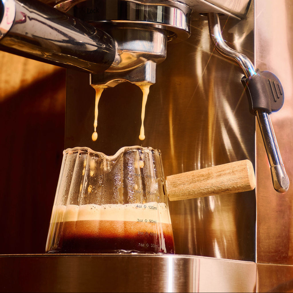 A close-up of a stainless steel espresso machine dispensing a stream of dark coffee with crema into a small glass cup with a wooden handle.