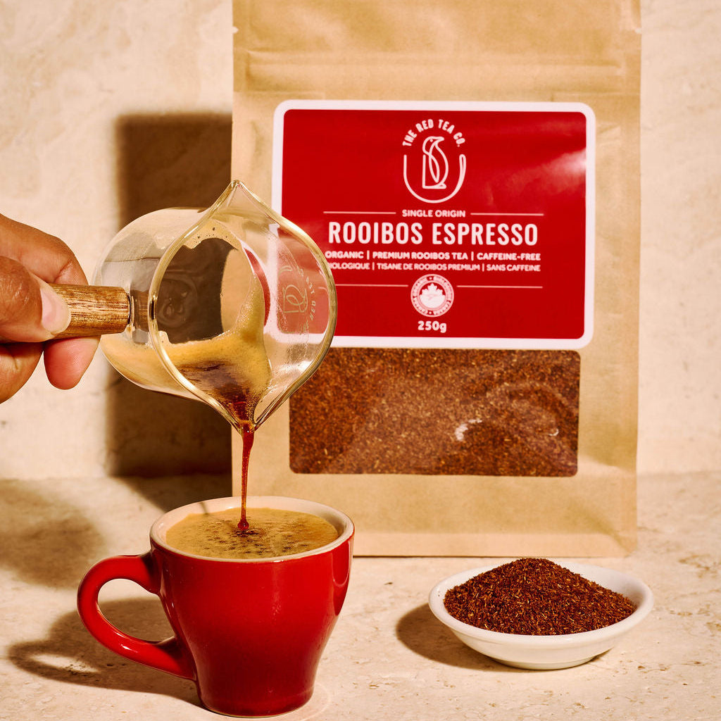 A hand pours dark rooibos espresso from a glass pitcher into a red mug, with a stand-up pouch of Rooibos Espresso tea and a small dish of loose powder behind it on a beige stone surface.